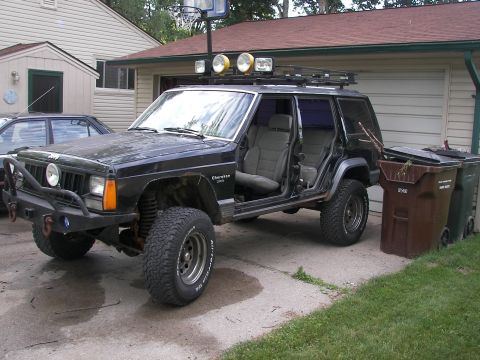 1988 Ford Bronco - This modified Jeep Cherokee XJ is parked in a driveway next to a house and trash cans.