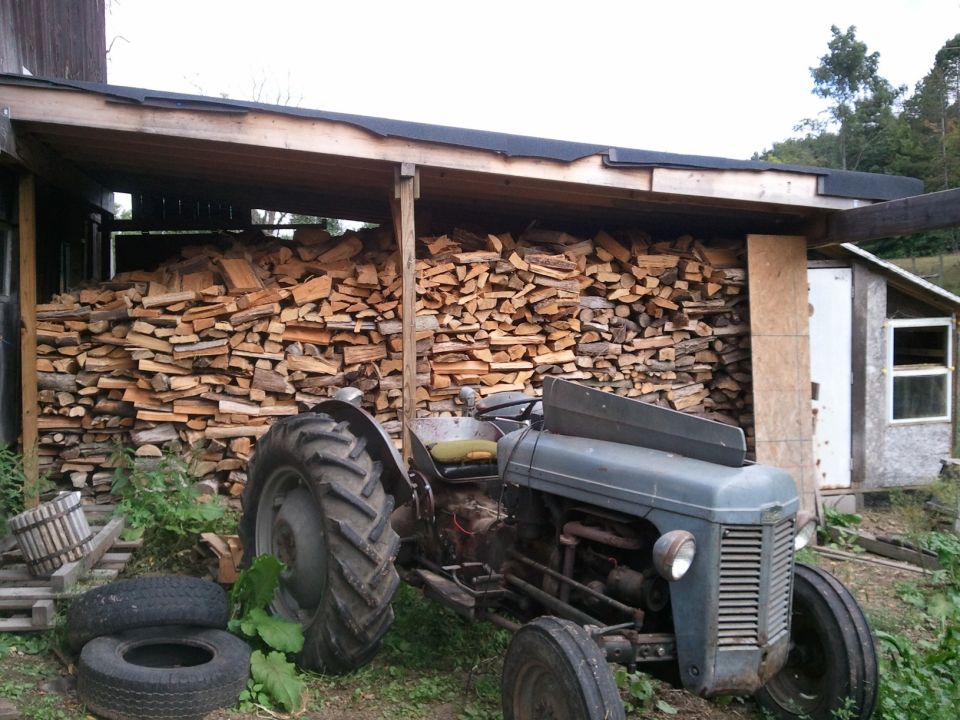 Old gray tractor parked in front of a woodpile next to a shed