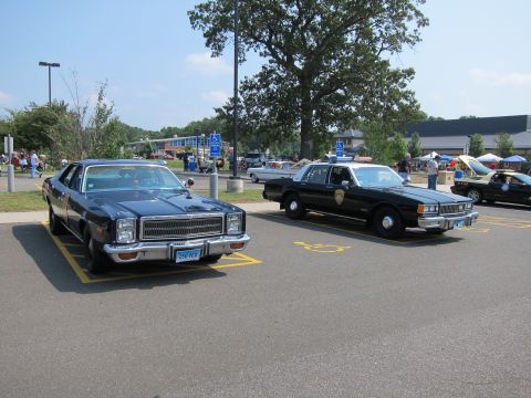 1991 Ford LTD Crown Victoria - Two classic cars in a sunny parking lot: a dark muscle-style sedan beside a black-and-white police cruiser.