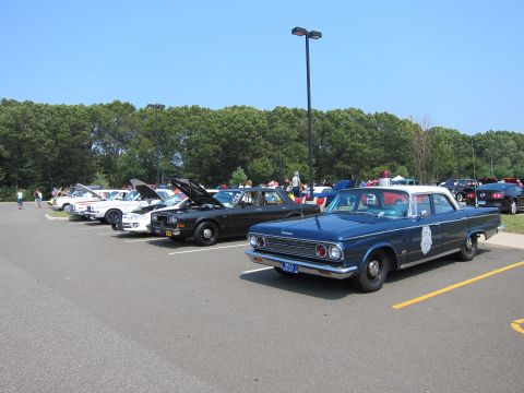 1991 Ford LTD Crown Victoria - blue 1960s-style police sedan with a door shield decal heads a row of classic vehicles in a sunny parking lot.