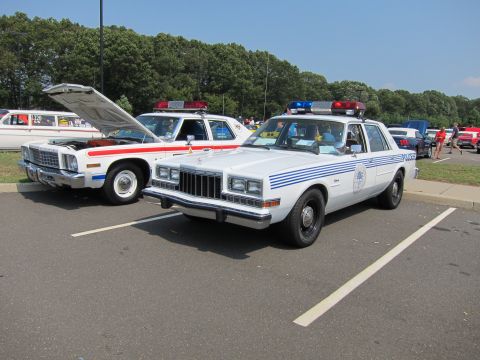 1991 Ford LTD Crown Victoria - white police sedan with blue striping, door shield and roof light bar parked beside another classic cruiser.