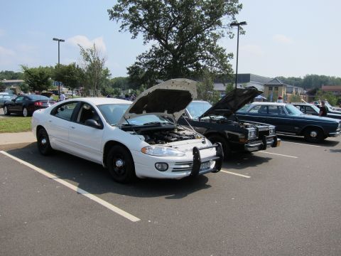 1991 Ford LTD Crown Victoria - white modern police-style sedan with its hood open parked beside a dark sedan and classic cars in a sunny lot.
