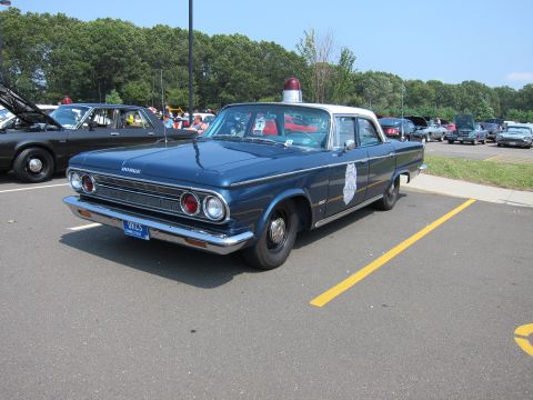 1991 Ford LTD Crown Victoria - blue 1960s-style police sedan with door shield, roof light and stacked headlights parked at a sunny car show.