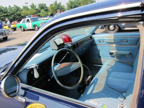 1991 Ford LTD Crown Victoria - Interior view through the open door of a vintage police car showing a blue bench seat, steering wheel and dash-mounted light.