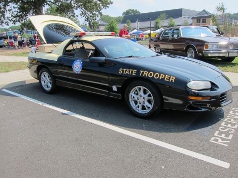 1991 Ford LTD Crown Victoria - black late-model Camaro lettered STATE TROOPER in gold sits with its hood up in a reserved show parking space.