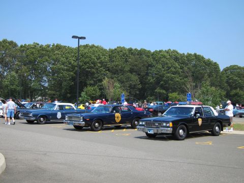 1991 Ford LTD Crown Victoria - row of dark-colored classic police cars with agency seals is displayed at a busy outdoor car show under a clear sky.