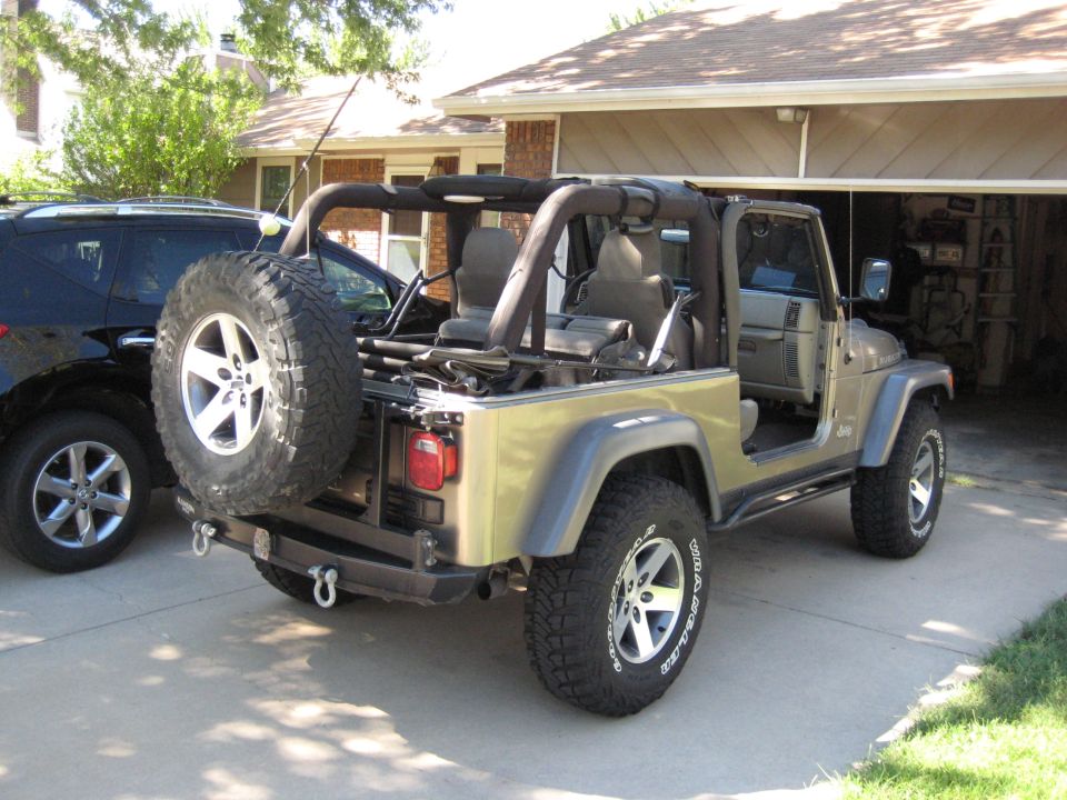 Tan Jeep Wrangler with spare tire on rear, parked in driveway next to black SUV