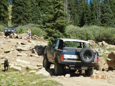 1996 Ford Bronco - Two off-road vehicles navigate a rocky mountain path with trees in the background.