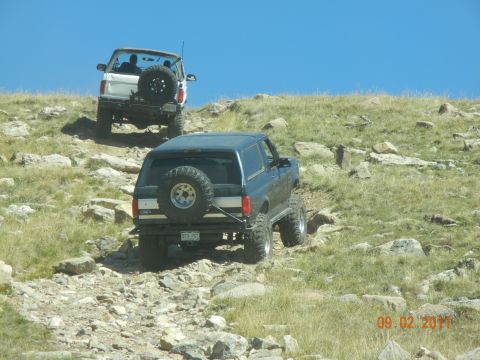 1996 Ford Bronco - Two SUVs navigate a steep, rocky trail under a clear blue sky. The lead vehicle is a dark SUV, followed by a white SUV.