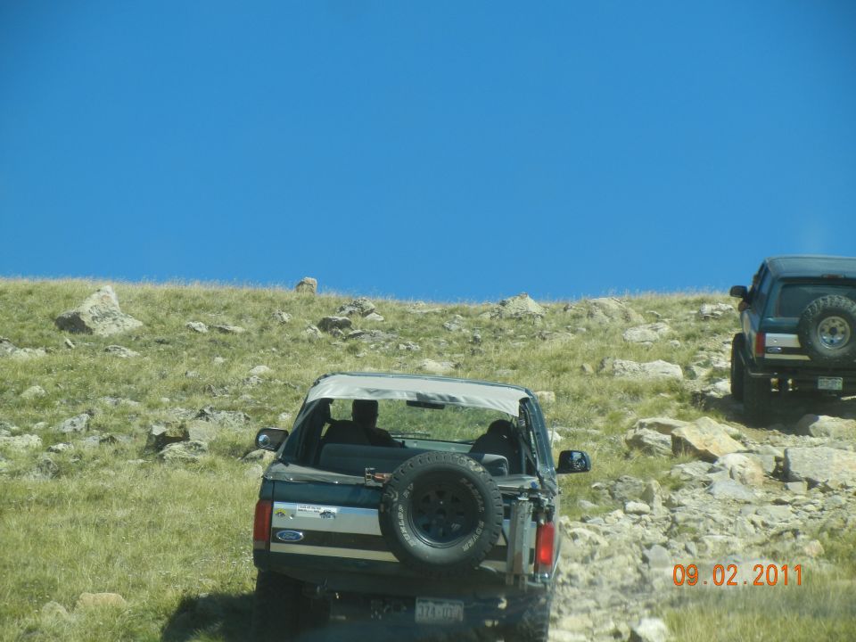 1996 Ford Bronco with spare tire on back, driving on a rocky, grassy slope under a clear blue sky. Another SUV is visible further up the hill