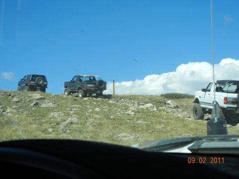 1996 Ford Bronco - Multiple SUVs traverse a , grassy incline under a bright blue sky with dramatic clouds.