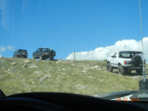 1996 Ford Bronco - Three SUVs, including a white 1996 Ford Bronco, navigate a rocky, grassy incline under a bright blue sky.