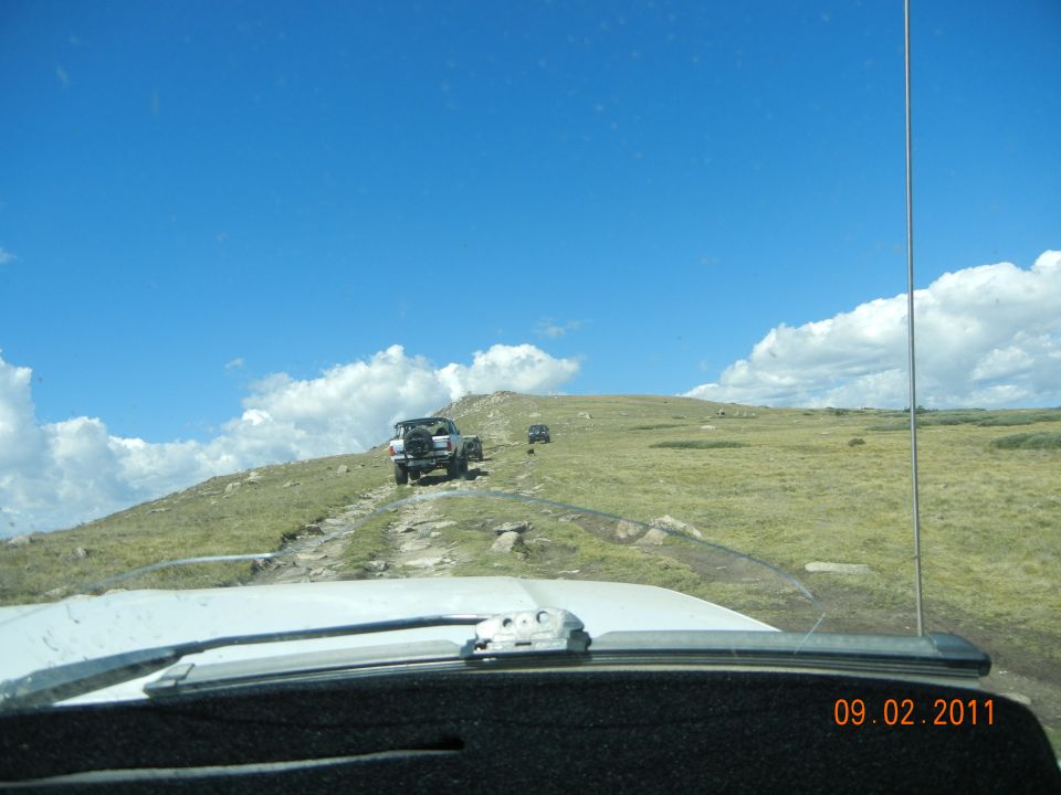 Several SUVs driving up a rocky dirt road on a grassy hill under a blue sky with clouds