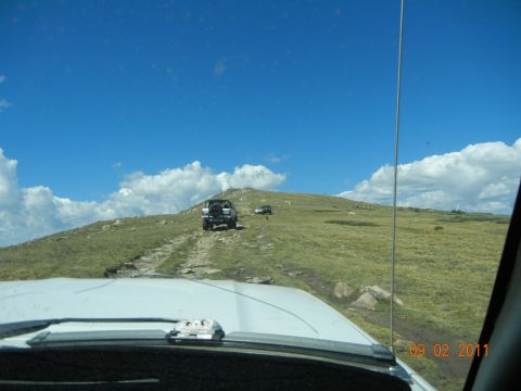 1996 Ford Bronco - Two SUVs navigate a rocky path on a mountain trail under a bright blue sky with scattered clouds.