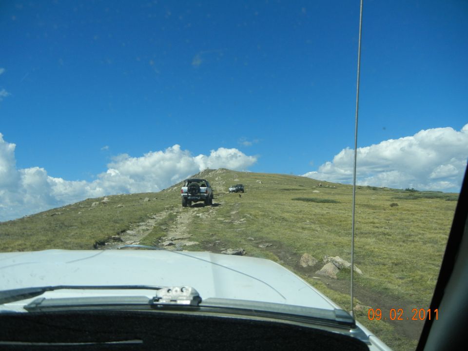 White Ford Bronco driving on a dirt road up a grassy hill under a blue sky with clouds