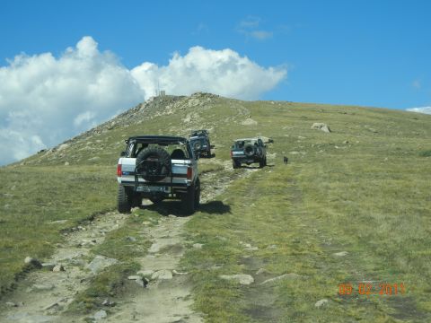 1996 Ford Bronco - Three Ford Broncos ascend a grassy, rocky mountain path under a blue sky with scattered clouds.