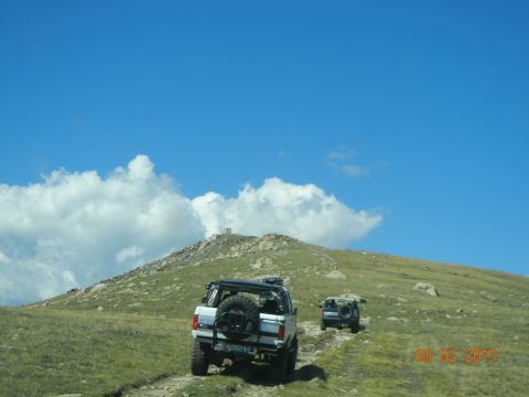 1996 Ford Bronco - Two SUVs navigate a dirt road on a grassy mountain slope under a bright blue sky with scattered clouds.