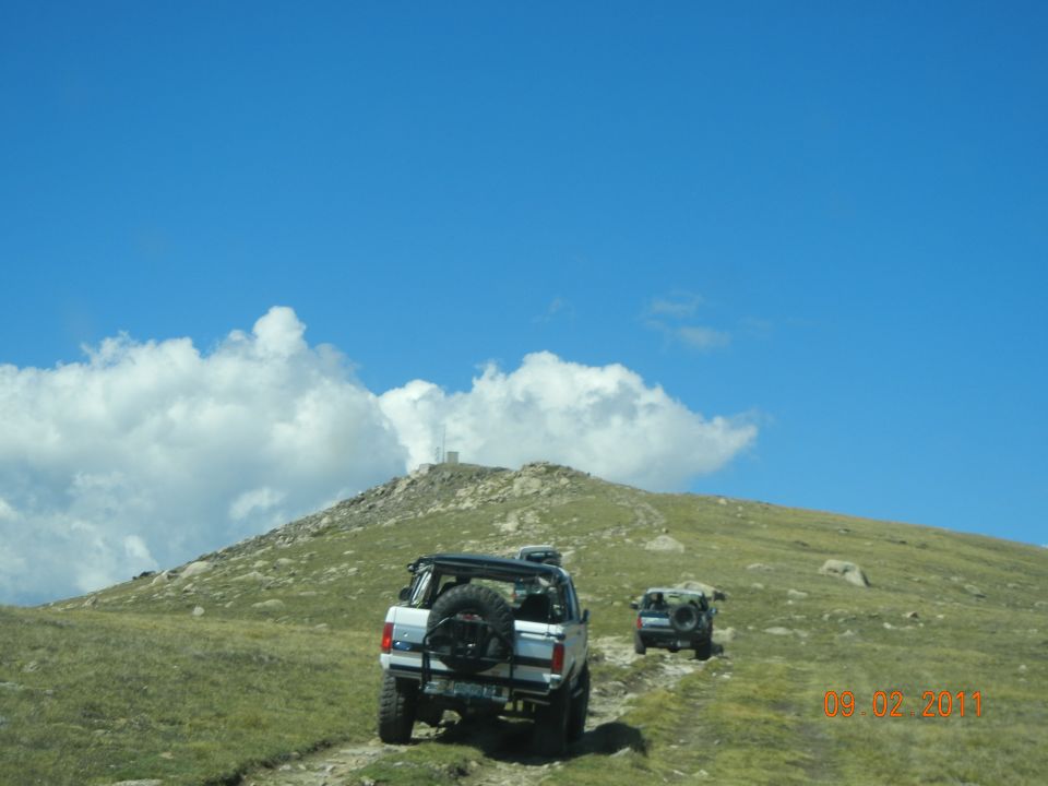 White SUV with spare tire on rear, driving on dirt road up grassy hill with another vehicle behind