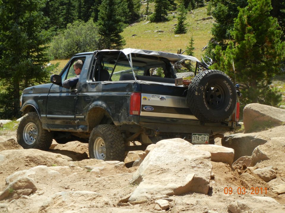 Black Ford Bronco with a soft top, lifted suspension, and a spare tire on the rear, navigating rocky terrain
