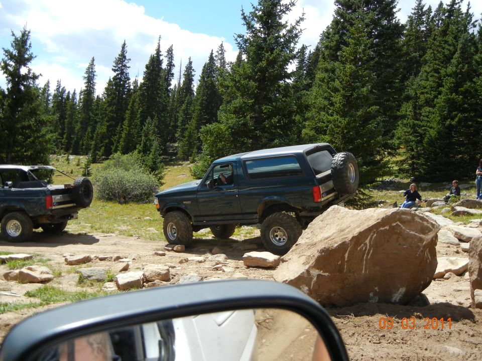 Dark green 1996 Ford Bronco climbing over a large rock on a dirt trail with trees in the background