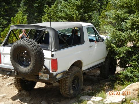 1996 Ford Bronco - A white 1996 Ford Bronco convertible navigates a rocky trail surrounded by trees.