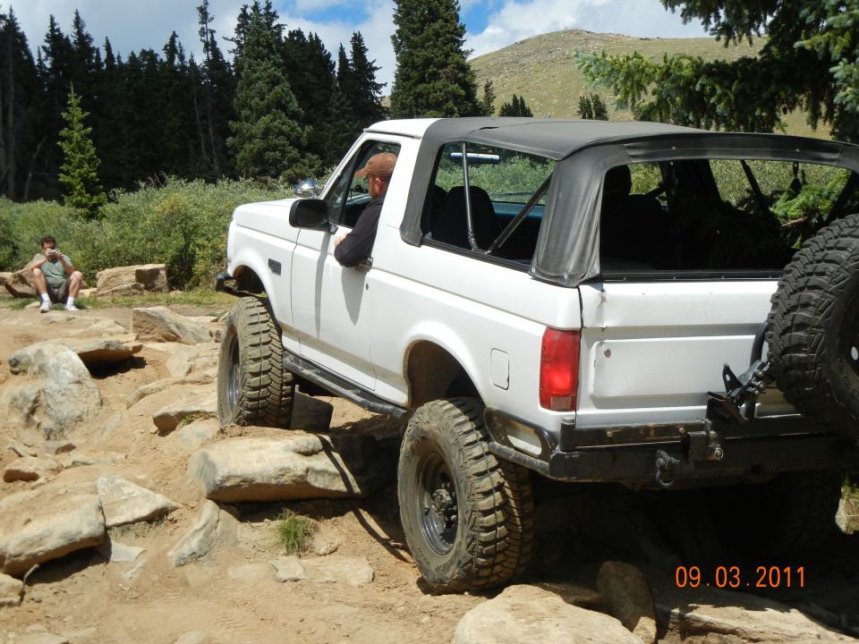 White 1996 Ford Bronco with soft top navigating a rocky off-road trail. Driver visible. Spare tire mounted on rear