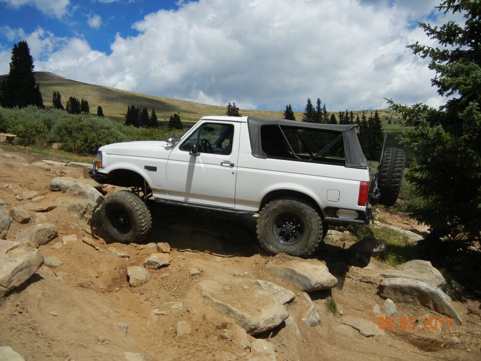 White 1992 Ford Bronco with a soft top, lifted suspension, and off-road tires navigating a rocky trail