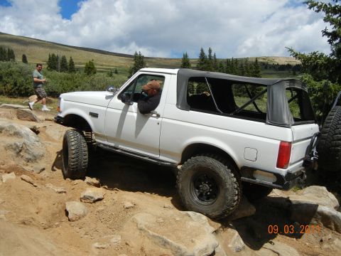 1996 Ford Bronco - A white SUV navigates a rocky off-road trail under a partly cloudy sky.