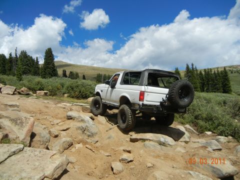 1996 Ford Bronco - A white 1996 Ford Bronco navigates a challenging rocky terrain under a cloudy blue sky.