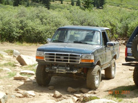 1996 Ford Bronco - A dark blue 1995 Ford Bronco navigates a rocky off-road trail, equipped with a winch and large tires.