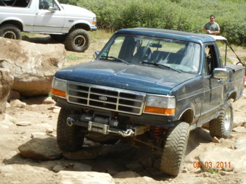 1996 Ford Bronco - A man watches as a dark blue Ford pickup truck navigates a rocky trail. Another Ford vehicle is visible in the background.