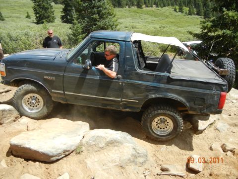 1996 Ford Bronco - A dark blue Ford Bronco navigates a rocky trail with a white soft top and spare tire mounted at the rear.