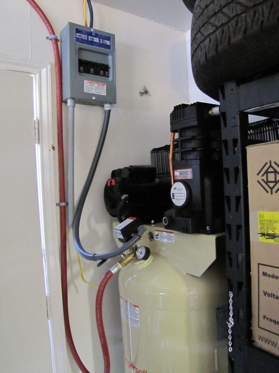Air compressor with gauge and electrical panel on wall. Tire on shelf above