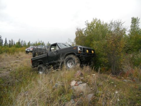 1986 Ford F-150 - Several trucks navigate a grassy, rocky off-road path with trees and foliage.