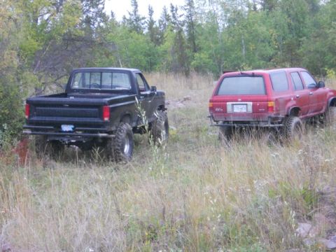 1986 Ford F-150 - Two off-road vehicles, a black pickup and a red SUV, are parked in a field with tall grass and trees in the background.