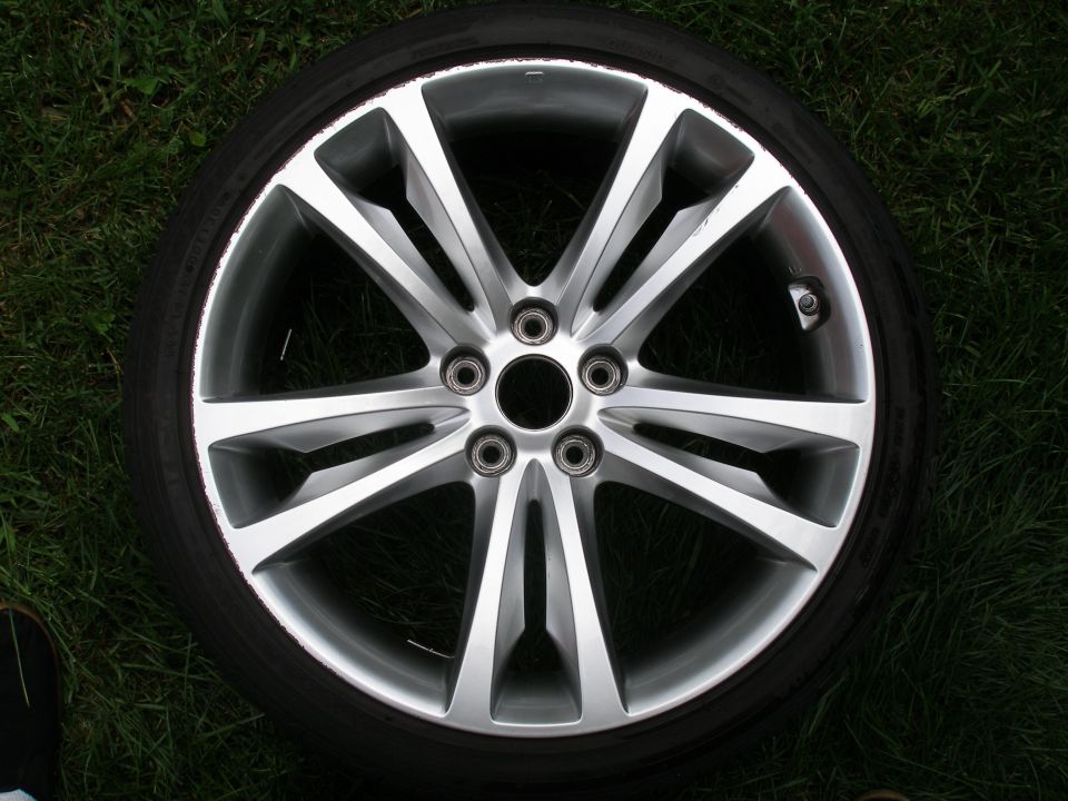 Close-up of a silver alloy wheel with a black tire, resting on grass. Supermotors.net logo in corner