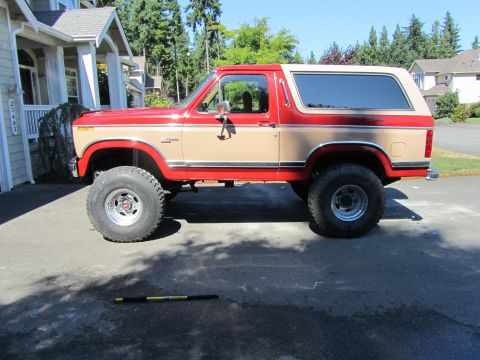1984 Ford Bronco - This two-tone red and tan Ford Bronco features a lifted suspension and oversized tires, suggesting off-road modifications.