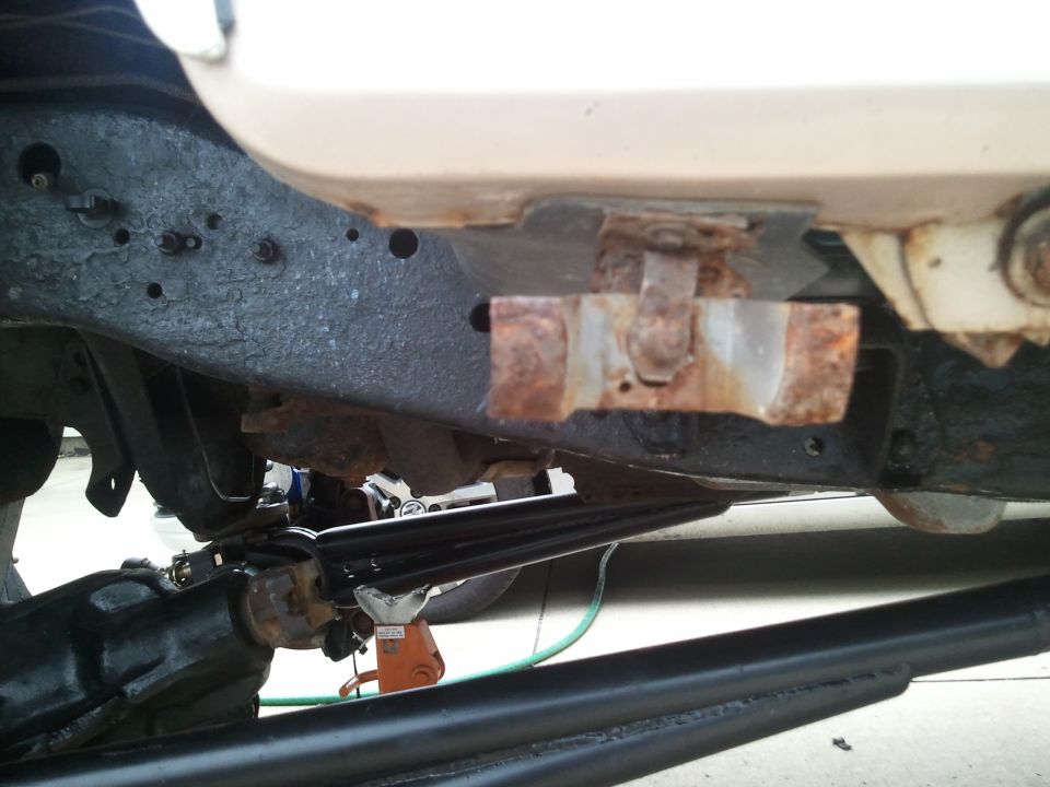 Underside of a vehicle frame with visible rust and suspension parts, including a drive shaft and orange jack