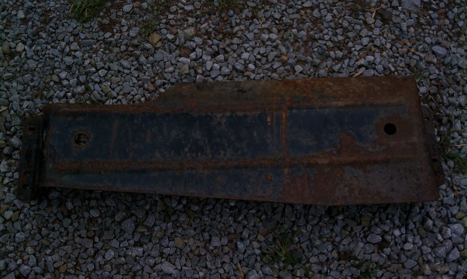 Rusty long steel bracket or skid plate with mounting holes lying on gray gravel