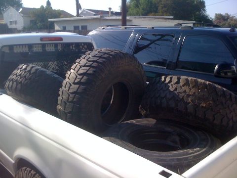 1987 Suzuki Samurai - Several large, knobby tires are piled in the bed of a white pickup truck, with a dark SUV visible behind.