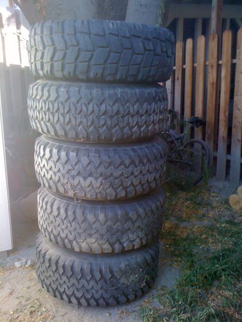 1987 Suzuki Samurai - A stack of five off-road tires are piled up next to a wooden fence with a bicycle in the background.