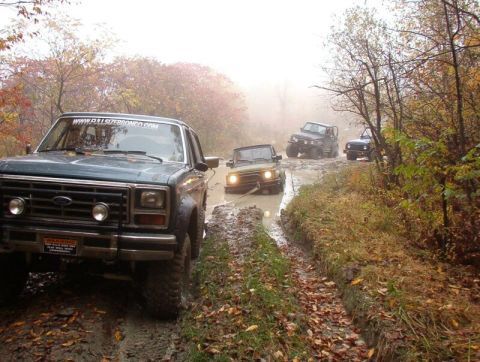 1986 Ford Bronco - A group of SUVs and off-road vehicles are stuck in a muddy trail during autumn, with trees and fog surrounding them.