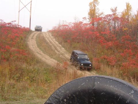 1986 Ford Bronco - Two off-road vehicles navigate a muddy track through autumn foliage.
