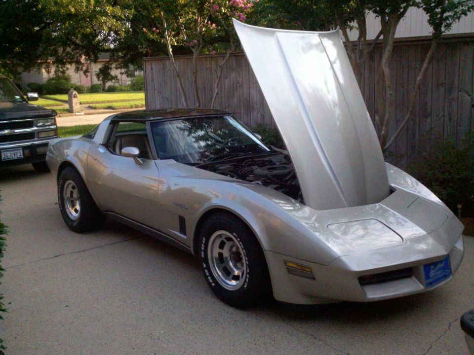 Silver 1982 Chevrolet Corvette with hood open, parked on a driveway next to a black pickup truck