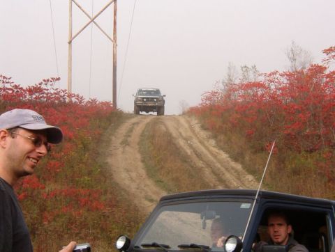 1986 Ford Bronco - Two SUVs navigate a dusty track surrounded by autumn foliage, with a utility tower in the background.