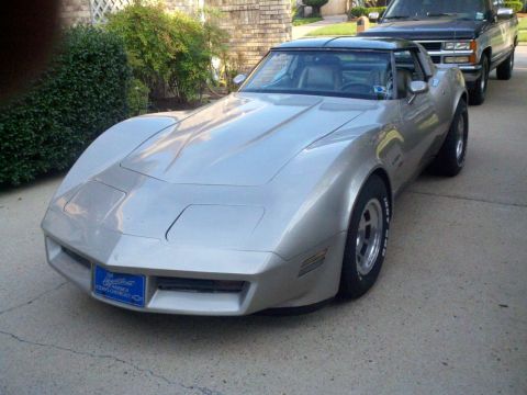 1982 Chevrolet Corvette - A silver 1982 Chevrolet Corvette is parked in front of a brick house, with a black pickup truck behind it.