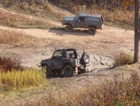 1986 Ford Bronco - Two off-road vehicles, a 1986 Ford Bronco and a Suzuki Samurai, navigate a muddy track.