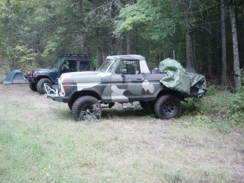 1979 Ford Bronco - Two off-road vehicles and a tent are set up in a clearing surrounded by trees.