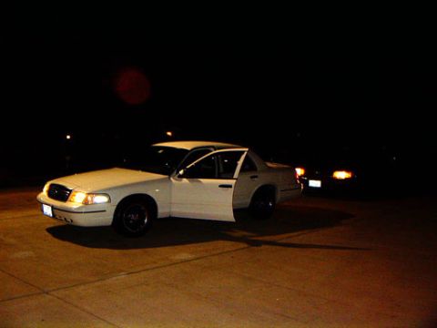 1999 Ford Crown Victoria - A white sedan is parked at night with its driver's door ajar, illuminated by its own headlights.