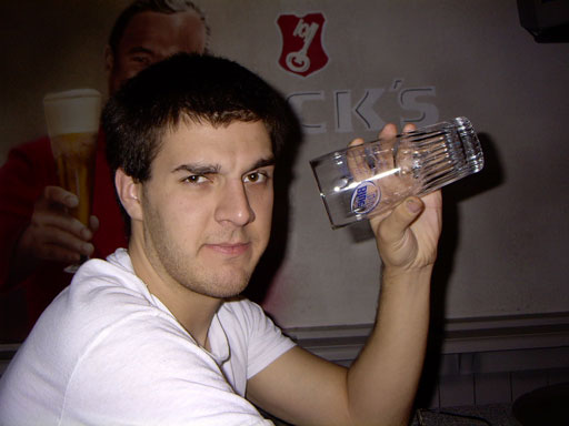 Man holds a clear beer glass with a blue label. Background shows a poster of a man holding a beer